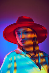 Trendy and artistic, image captures female model in a bold red wide-brimmed hat and satin shirt against gradient purple background in neon light. Concept of modern fashion, trendy style, beauty, youth
