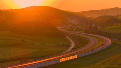AERIAL, LENS FLARE: Sun slowly setting over a winding road and scenic landscape. Evening sunbeams...