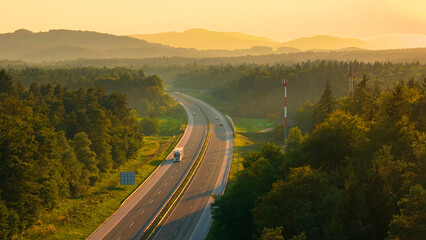 Obraz premium AERIAL: Trucks and cars travel along the scenic highway in golden evening light. Asphalt motorway winds through the beautiful wooded and hilly countryside, bathed in the rays of setting summer sun.
