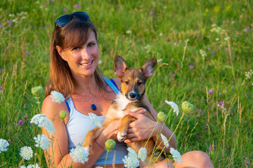 PORTRAIT, DOF: Blooming meadow with a pretty lady posing with her young puppy. Smiling woman lovingly holds her small puppy amidst a field of wildflowers. Moments of happiness with a furry companion.