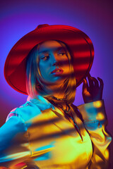 Trendy and artistic, image captures female model in a bold red wide-brimmed hat and satin shirt on gradient purple background in neon. Concept of modern fashion, trendy style, beauty, youth culture