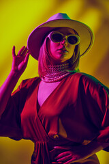 Contemporary style image with female model in fedora hat and layers of pearl necklaces, red dress against yellow background in neon light. Concept of modern fashion, trendy style, beauty, youth