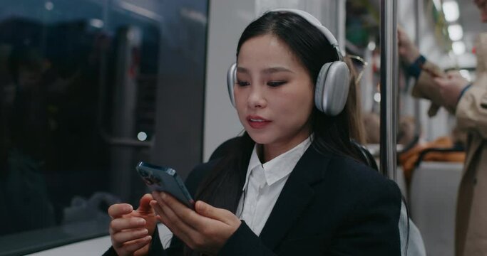 Chinese girl coming back home from work or university in bus or train. Wearing black and white uniform. Enjoying music in large headphones. Taking out phone to scroll social media or world news.