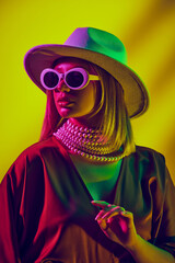 Retro vibes and modern style, hat, sunglasses, pearl neckless and dress. Model posing against yellow background in neon light. Concept of modern fashion, trendy style, beauty, youth culture