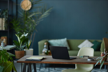 Background image of modern office interior with laptop on wooden desk and teal wall with green live plants copy space 
