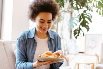 Beautiful smiling African American woman holding croissant while sitting on comfortable sofa in room