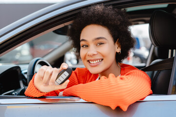 Smiling African American woman with curly hair sitting in car salon, happy owner holding car key