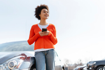Happy African American woman with curly hair holding mobile phone, standing near car, looking away