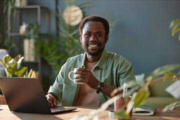 Portrait of smiling African American man looking at camera while sitting at workplace in office decorated with green live plants copy space 