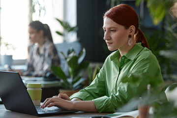 Sde view portrait of young red haired businesswoman using laptop at workplace in office decorated with live green plants copy space 
