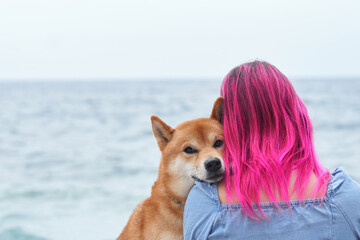 A Shiba Inu dog cozily nestles into a woman with striking pink hair, overlooking the tranquil sea....