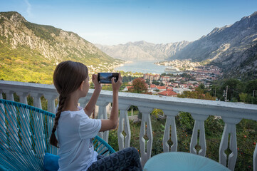 Rear view of little girl take pictures with her smartphone from the terrace of a room with an amazing panoramic view of the picturesque Bay of Kotor in Montenegro