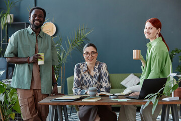 Front view portrait of multiethnic business team of three people smiling at camera in office against teal wall