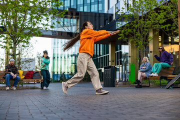 Young Stylish Female Hip-Hop Dancer Freestyling On Urban City Street Among Modern Buildings. Fashionable Friends Relaxing On Background, Supporting Street Style Performer Practising Choreography.