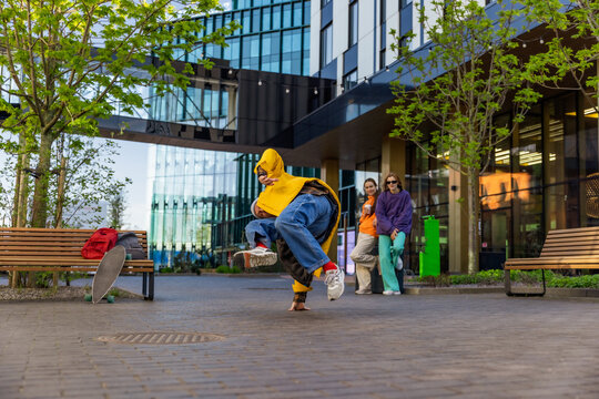 Young Stylish B-boy Breakdancing On City Street Among Modern Buildings In Urban Area. Fashionable Group Of Friends Relaxing On Background, Supporting Breakdance Performer Practising Choreography.