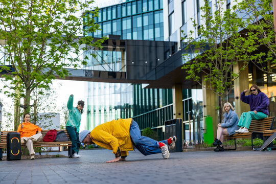 Young Stylish B-boy Breakdancing On City Street Among Buildings In Urban Area. Fashionable Group Of Friends Chilling On Background, Supporting Breakdance Performer Practising Choreography.