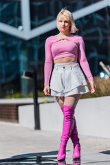Vertical Screen. Striking Image Of A Young Blonde Woman In A Pink Crop Top And White Skirt, Paired With Pink Boots, Walking Confidently In An Urban Setting With Modern Architecture In The Background. - obrazy, fototapety, plakaty