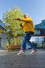 Vertical Screen: A Vibrant Young Man In A Yellow Jacket And Jeans Dances Energetically In An Urban Park, Showcasing A Breakdance Move Under A Blue Sky, Expressing Freedom And Joy In A City Setting.
