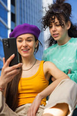 Two Young Women Enjoying Time Together Outdoors With a Smartphone, Captured In a Vertical Screen Format. One Wears a Purple Beret, the Other a Green Sweatshirt, Both Smiling, With Urban Background.