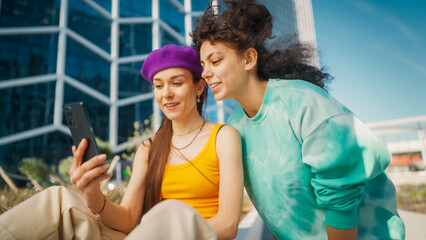 Two Young Women, One Wearing A Purple Hat And Yellow Top, The Other In A Green Sweatshirt, Share A Joyful Moment Looking At A Smartphone Together In An Urban Setting With A Glass Building Behind.