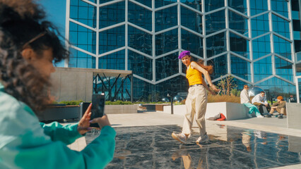 Vibrant Urban Dance Scene With A Young Woman In A Purple Beanie And Yellow Top Performing A Dynamic Dance Move, Captured By A Friend On A Smartphone, Against A Modern Glass Building Background.
