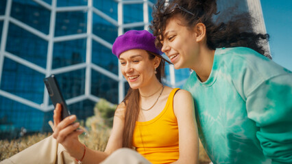 Two Joyful Young Women Sharing A Moment Over A Smartphone, With A Vibrant Cityscape Behind Them. They Are Dressed Casually, Emphasizing A Relaxed, Friendly Atmosphere Under The Bright, Sunny Sky.