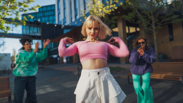 Outdoor Scene Captures A Young Blonde Woman Dancing Energetically In A Pink Top And White Skirt, With Friends Clapping And Capturing The Moment On A Smartphone, Embodying Youth In An Urban Setting.