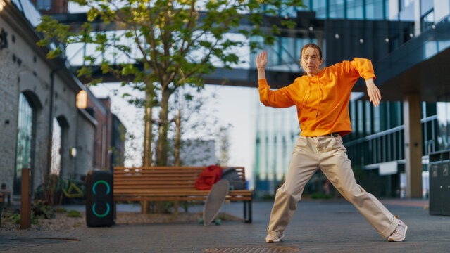 Urban Dance Scene With A Young Woman In An Orange Hoodie Performing Dynamic Moves On A City Street, With A Portable Speaker And Architecture In The Background. Youthful Energy And Street Culture.