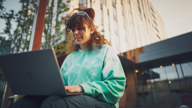 A Young Woman Enjoys The Outdoors While Working On Her Laptop, Showcasing A Blend Of Technology And Leisure In An Urban Setting, With A Modern Building In The Background.