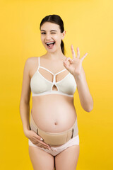 Portrait of pregnant woman in underwear wearing pregnancy bandage to make the backache go away and showing okay gesture at yellow background. Copy space. Orthopedic abdominal support belt concept