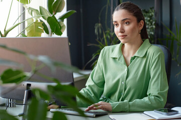 Portrait of smiling young businesswoman using computer sitting at workplace with green live plants copy space 