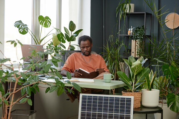 Portrait of adult African American man reading notes in planer while sitting at workplace with lush green plants copy space 