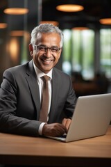 Smiling senior Indian business executive sitting at desk using laptop.