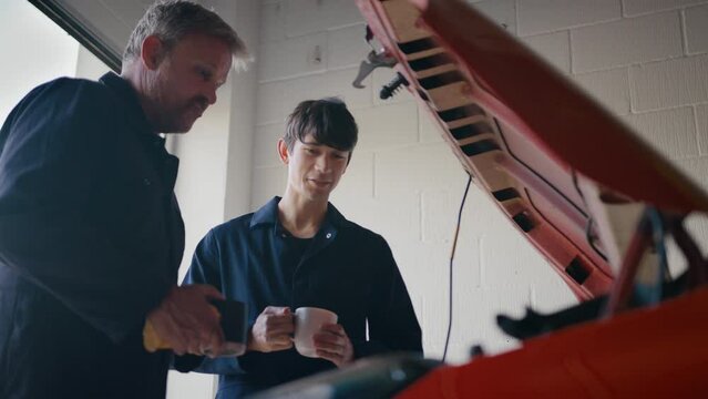 Experienced car mechanic servicing car engine looking under hood with male trainee drinking hot drink - shot in slow motion