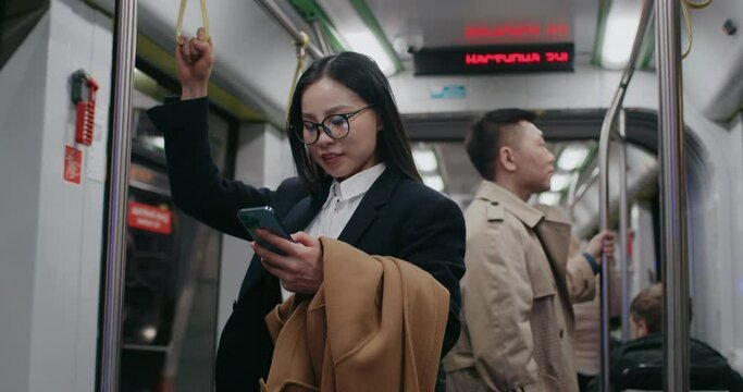 People traveling by metro or train. Going home in evening. Woman in front holding jacket with one hand while using smartphone. Glasses for better vision. Concept of public transportation.