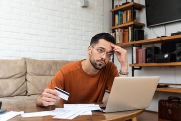Young stressed man having issue with utility bills expense, sitting at home trying to calculate and see bad finance bank report, searching for mistake. College student male with money problem.