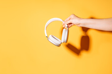 Hand holding white wireless headphones against a yellow background under hard light.