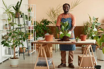 Full length portrait of smiling African American man repotting plants at home and enjoying...