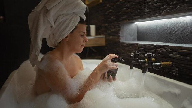 A serene woman with a towel on her head relaxes in a bubble bath, using soap indoors.