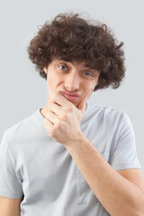 Funny and handsome, a young man with curly hair, he looks into the camera with his blue eyes, his hands on his face, a portrait of a guy wearing a T-shirt isolated against a gray background