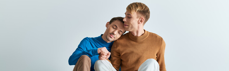 Two men in casual attires, one holding his leg up, share a moment of intimacy on a gray backdrop.