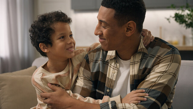 Smiling African American ethnic male man father dad using mobile phone device scrolling social media little boy son child kid running embrace hugging parent happy family indoors home living room couch - Powered by Adobe