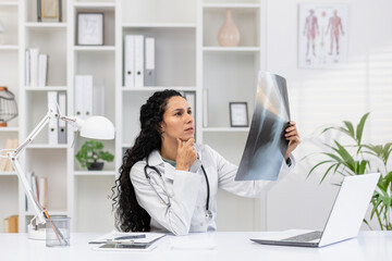 Focused Hispanic female doctor examining an X-ray film while contemplating diagnostics in a well-equipped modern medical office.