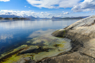 The serene sea, clear waters revealing submerged rocks, bordered by majestic mountains under a cloud-speckled sky.