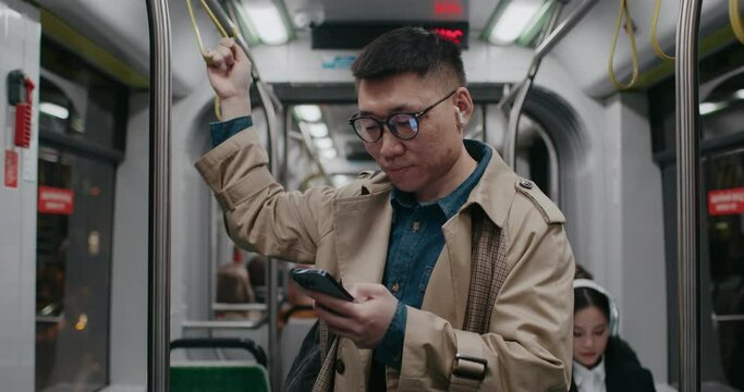 Stable front view of Asian male standing still in middle of long train wagon. Going back home after long working week. Browsing social media on smartphone. Listening to music in earphones.