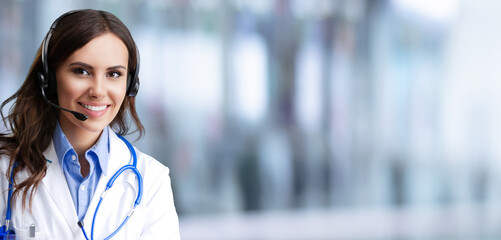 Portrait image of happy smiling female doctor in headset, against blurred modern clinic office background, blank mock empty area for ad slogan text. Medical call center. Wide banner