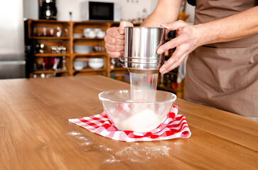 Person sifts flour into bowl on wooden table for recipe preparation