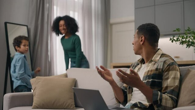 African American family quarrel busy annoyed male man father freelancer online working laptop notebook yell at dancing noisy mother female woman dance moves little child boy son kid leaving room house