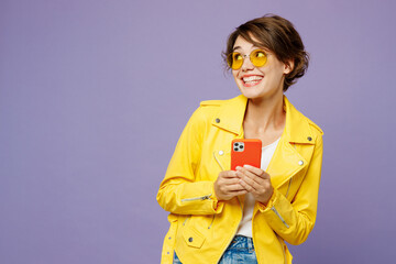 Young smiling woman wears yellow shirt white t-shirt casual clothes glasses hold in hand use mobile cell phone look aside on area isolated on plain pastel light purple background. Lifestyle concept.