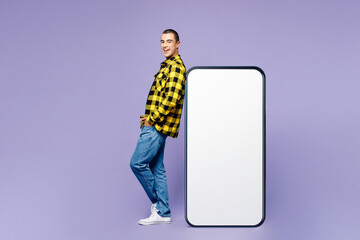 Full body side view young middle eastern man wears yellow shirt casual clothes big huge blank screen mobile cell phone smartphone with workspace area look camera isolated on plain purple background.